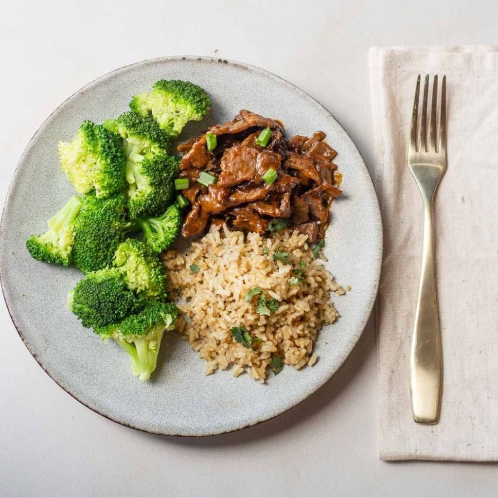 Plate with steamed broccoli, Mongolian beef, and brown rice, next to a gold fork on a beige napkin.