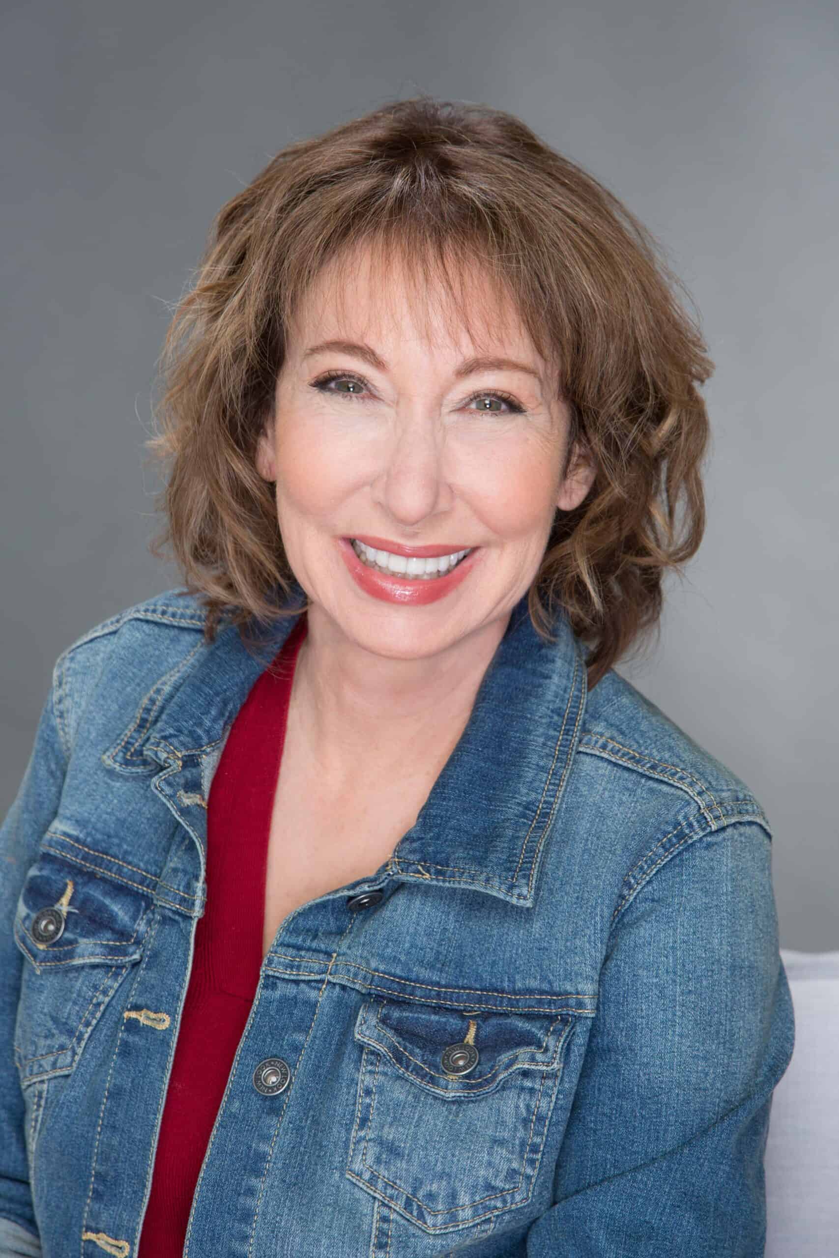 Smiling woman with short brown hair wearing a red top and denim jacket, against a gray studio backdrop.