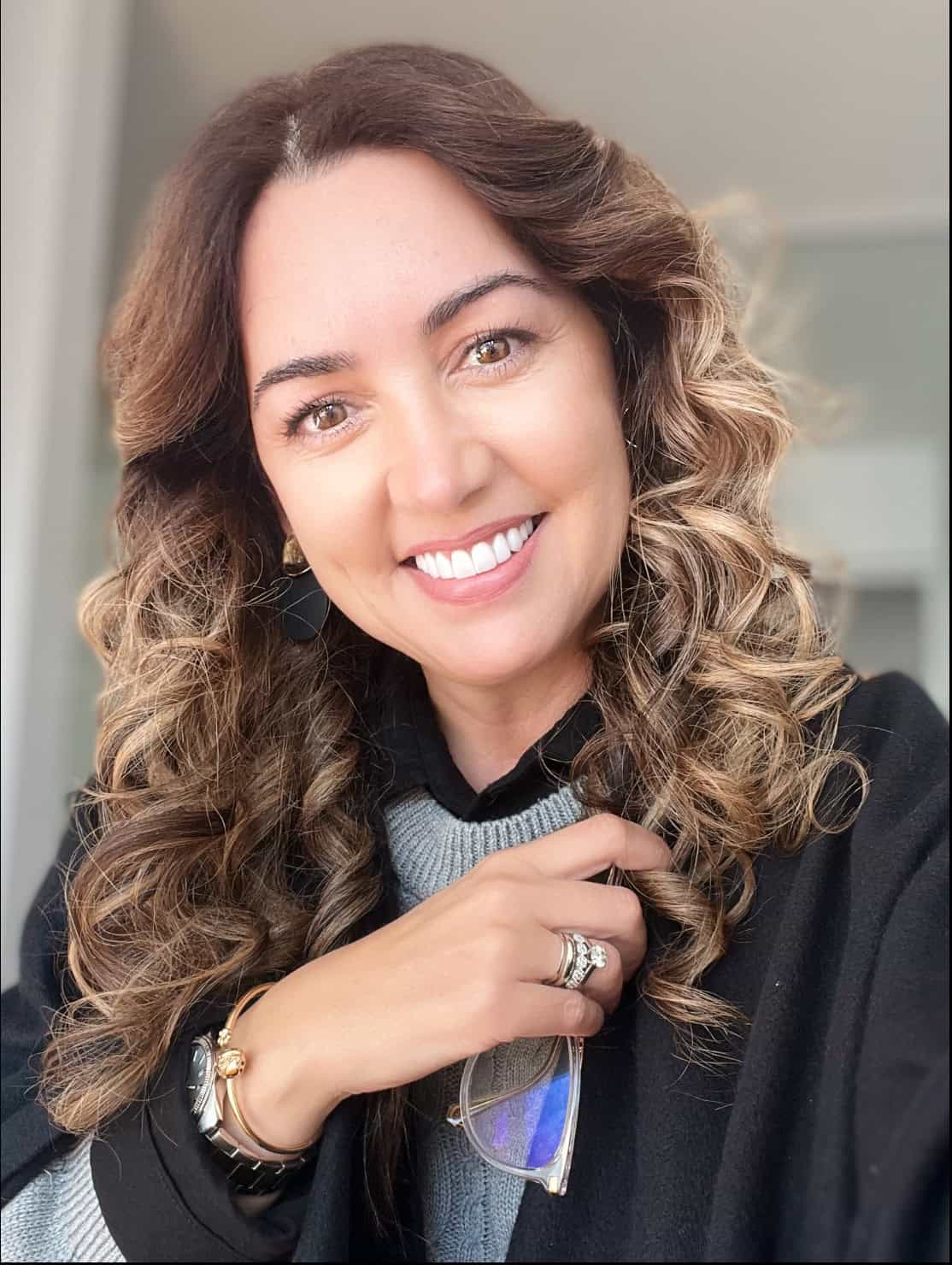 Smiling woman with wavy brown hair, wearing a gray sweater and black coat, holding sunglasses near her chest.