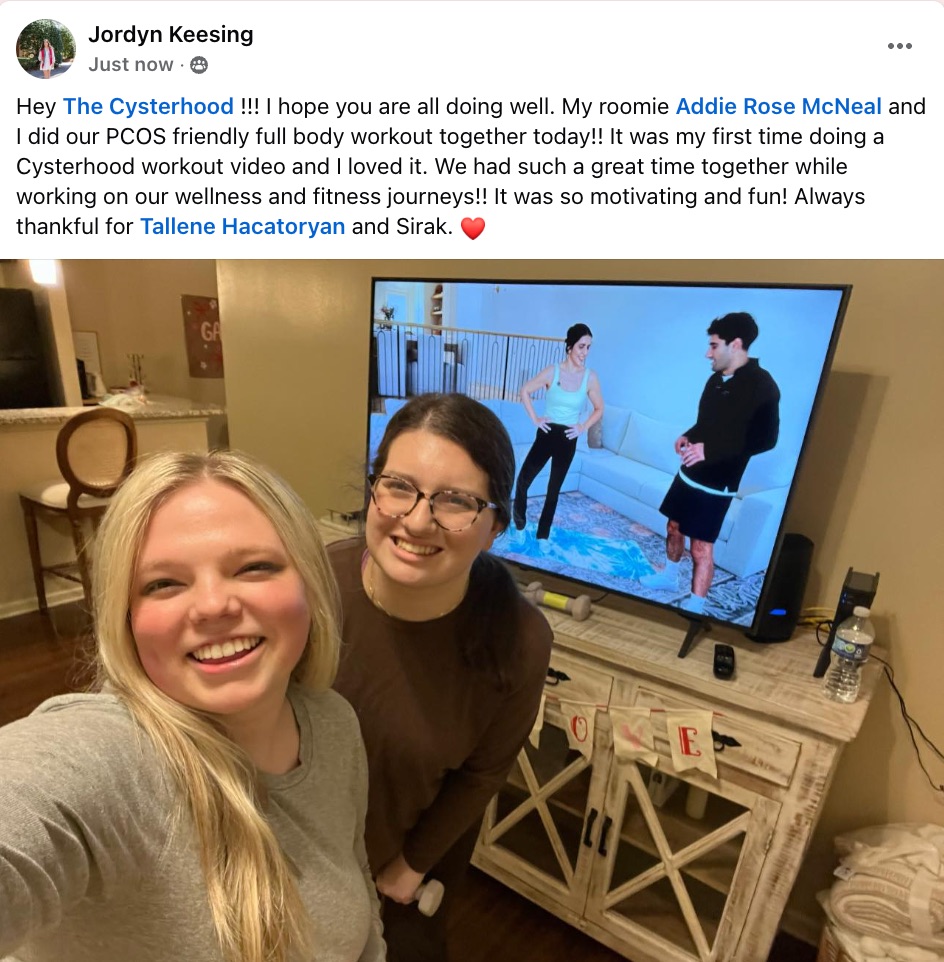Two smiling women take a selfie in a cozy living room as they follow a workout video on the TV. The screen shows a man and a woman exercising.