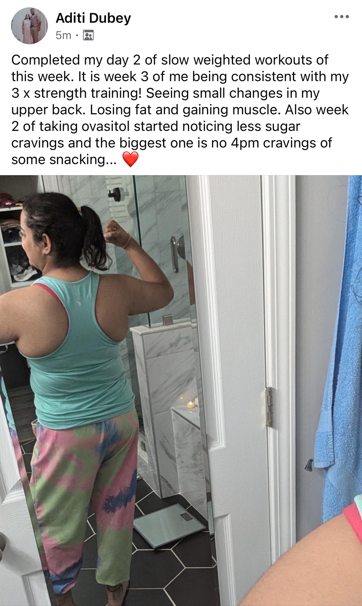 Back view of a woman flexing her bicep in a teal tank top and tie-dye pants, standing in a bathroom with a mirror.