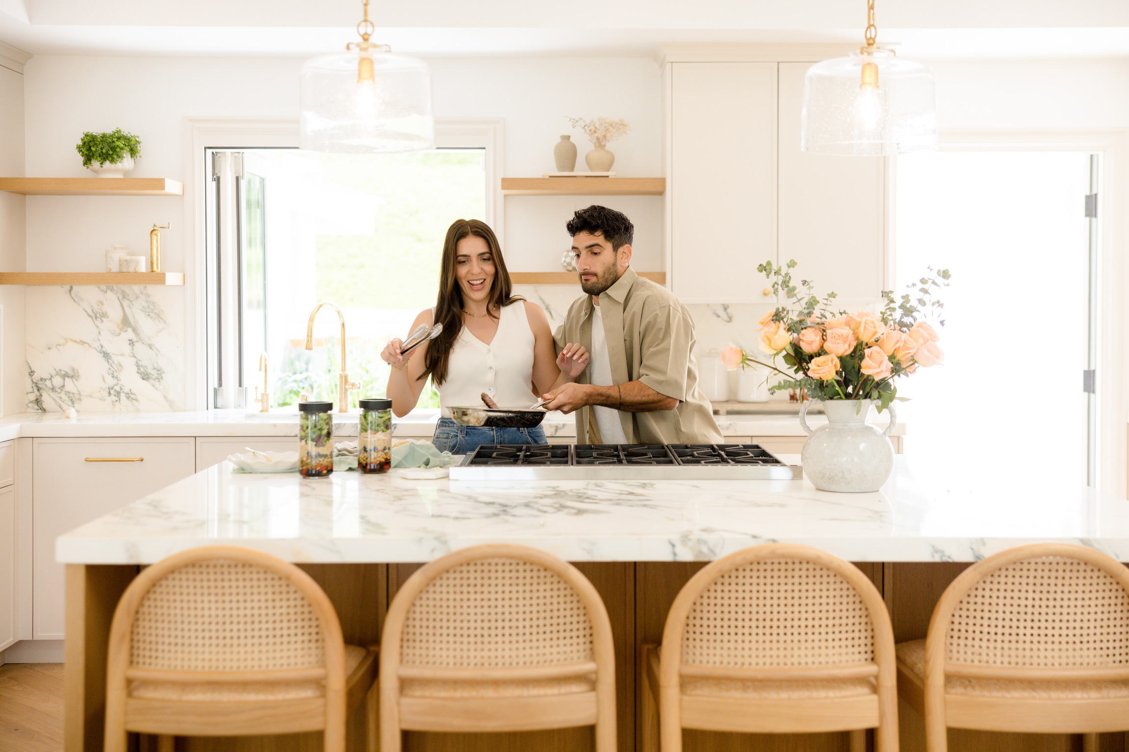 A couple cooks together at a bright marble kitchen island, with peach roses in a vase to the right and open shelves in the background.