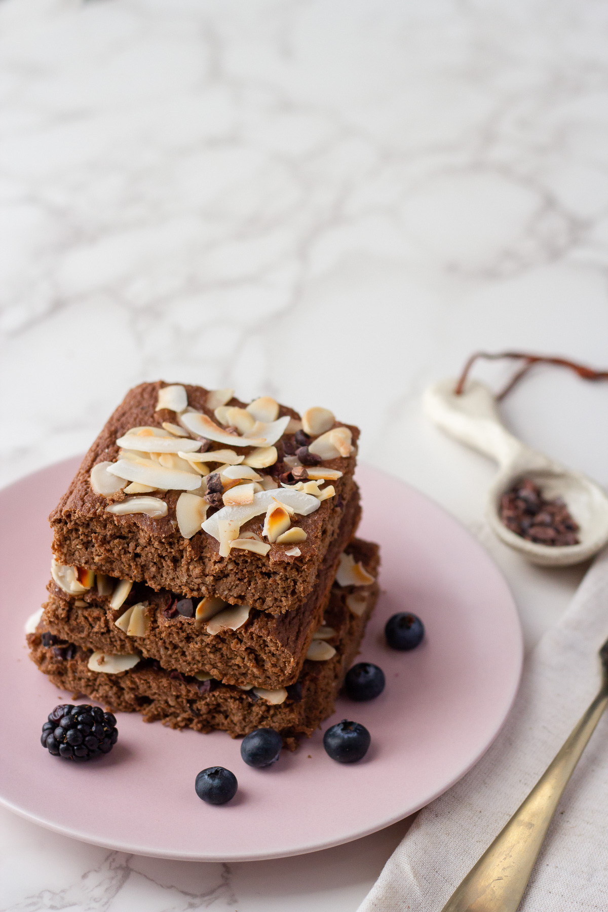 Stack of chocolate brownies topped with almond slices on a pink plate, with blueberries nearby on a marble surface.