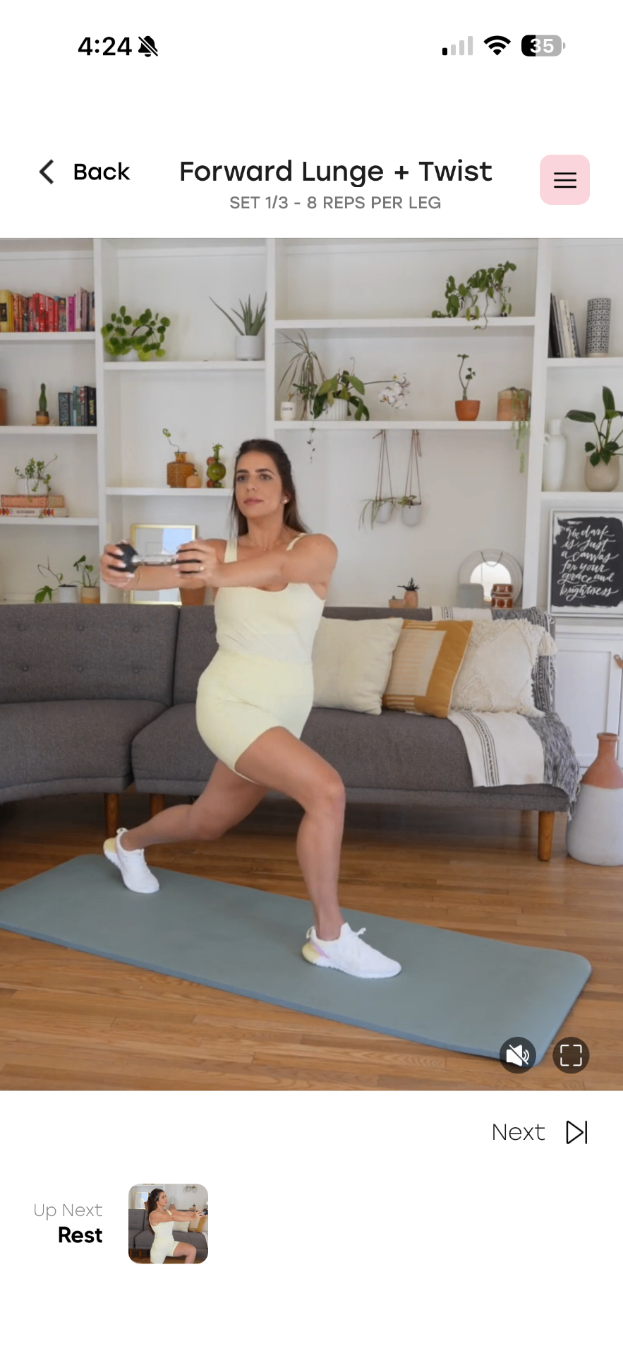 Woman in a pale yellow outfit performs a forward lunge with twist on a blue yoga mat in a living room filled with shelves and plants.