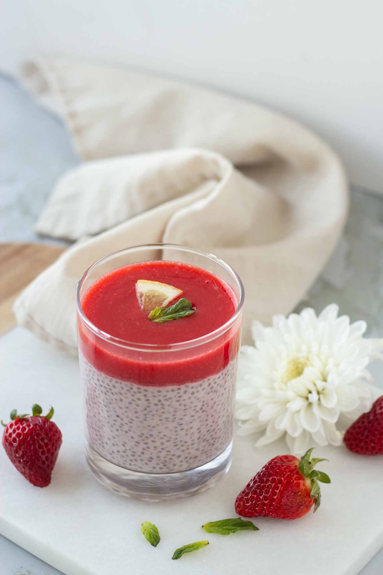 Layered berry smoothie in a glass with a chia-seed base, garnished with a lemon wedge and mint, on a white marble surface with strawberries nearby.