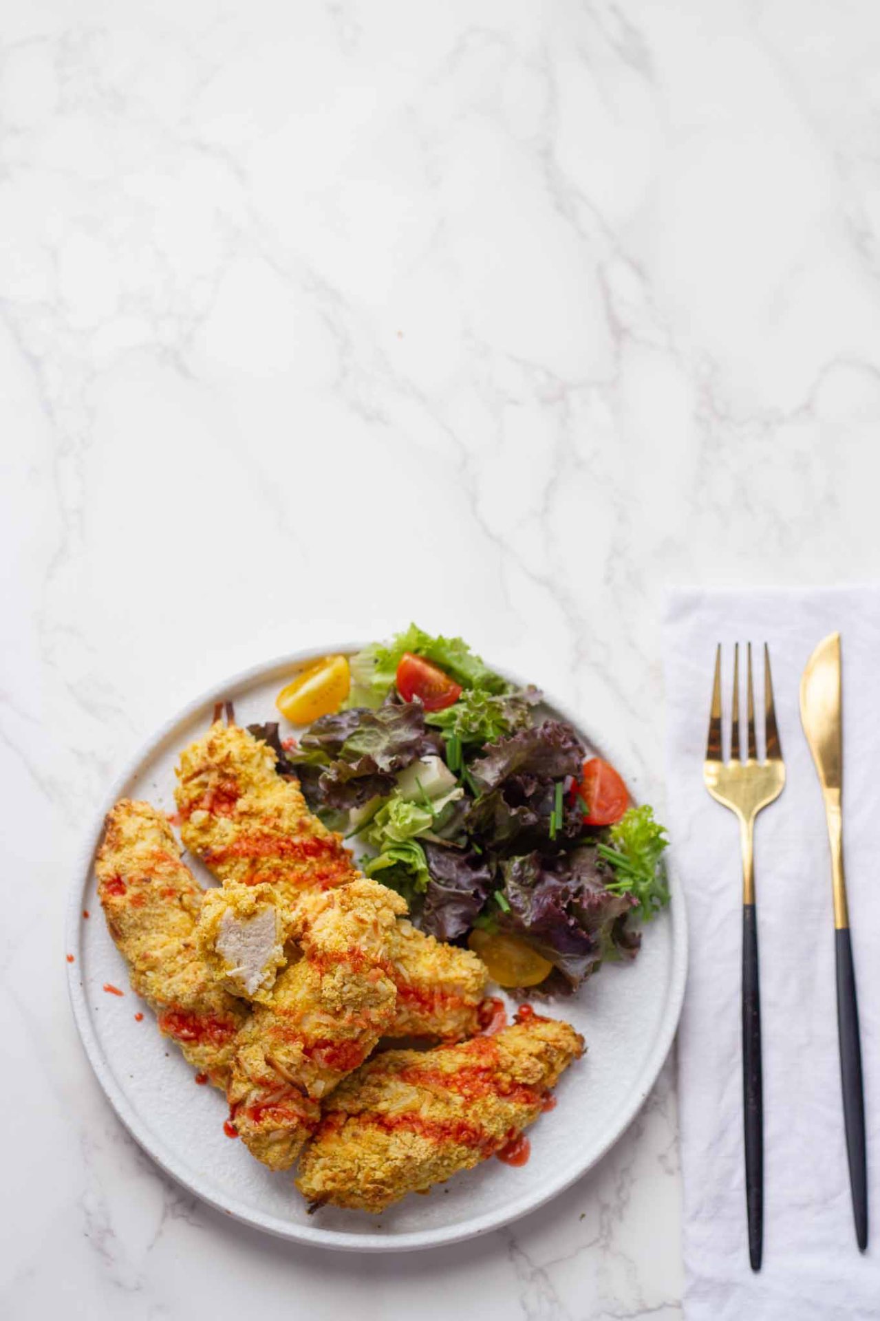 Plate of crispy fried chicken with a side salad of greens and tomatoes on a white marble table, with gold-edged cutlery nearby.
