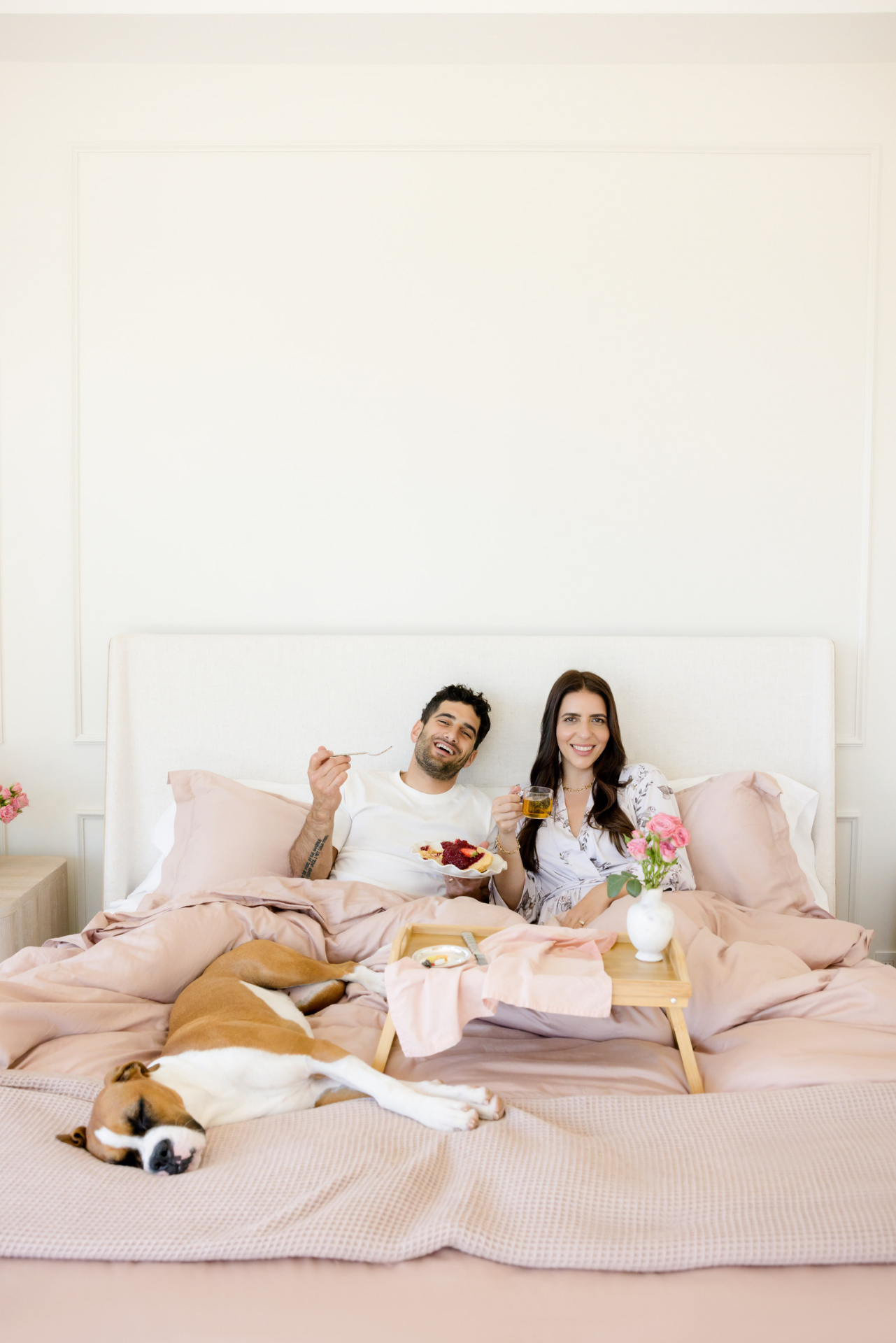 Couple in bed with a breakfast tray, smiling, as a dog sleeps at the foot of the bed.