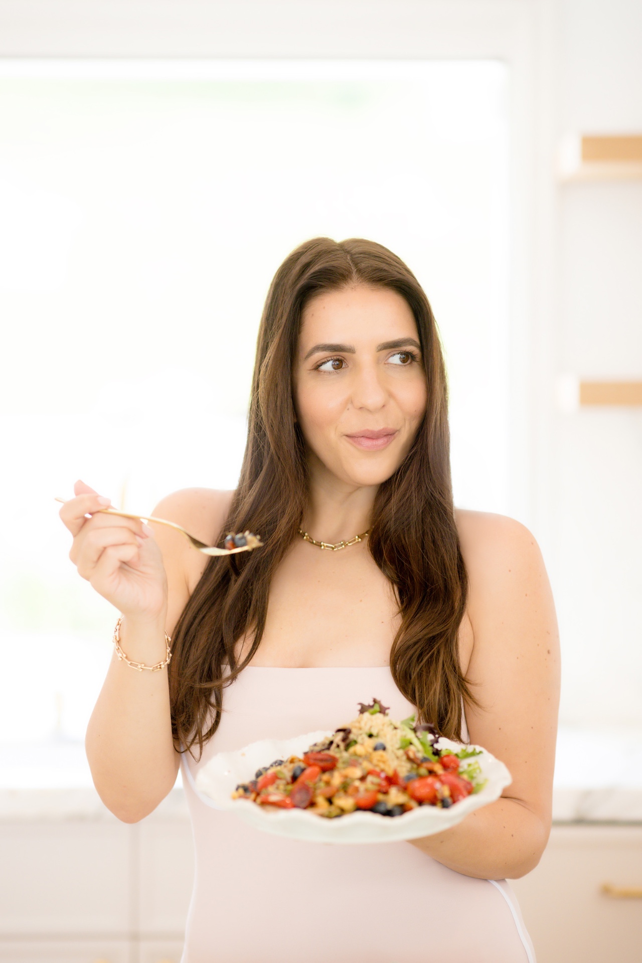 Woman in a white outfit holding a plate of colorful salad with a fork in a bright kitchen, smiling slightly to the side of camera.
