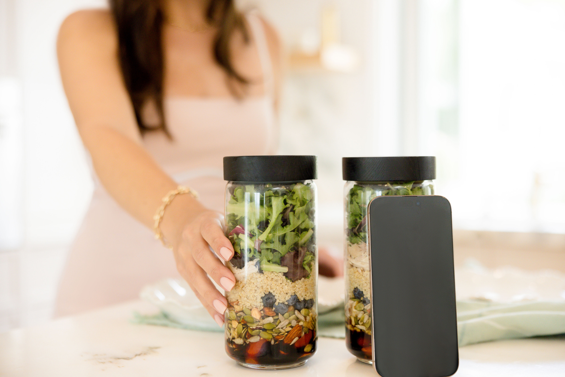 Hand reaching for a jar of layered salad on a kitchen counter with a smartphone nearby.