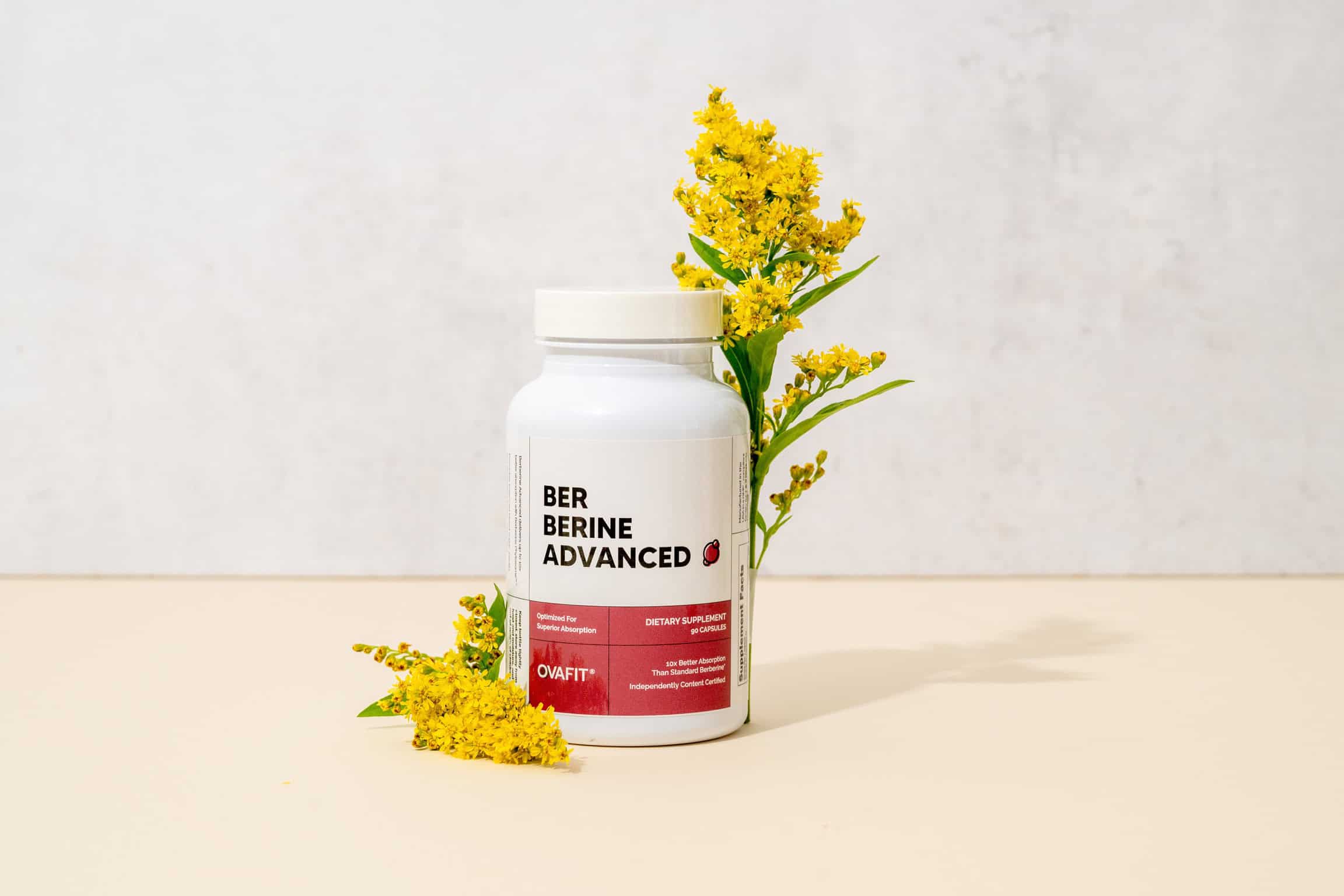White dietary supplement bottle labeled Berberine Advanced with yellow flowers beside it on a light background.