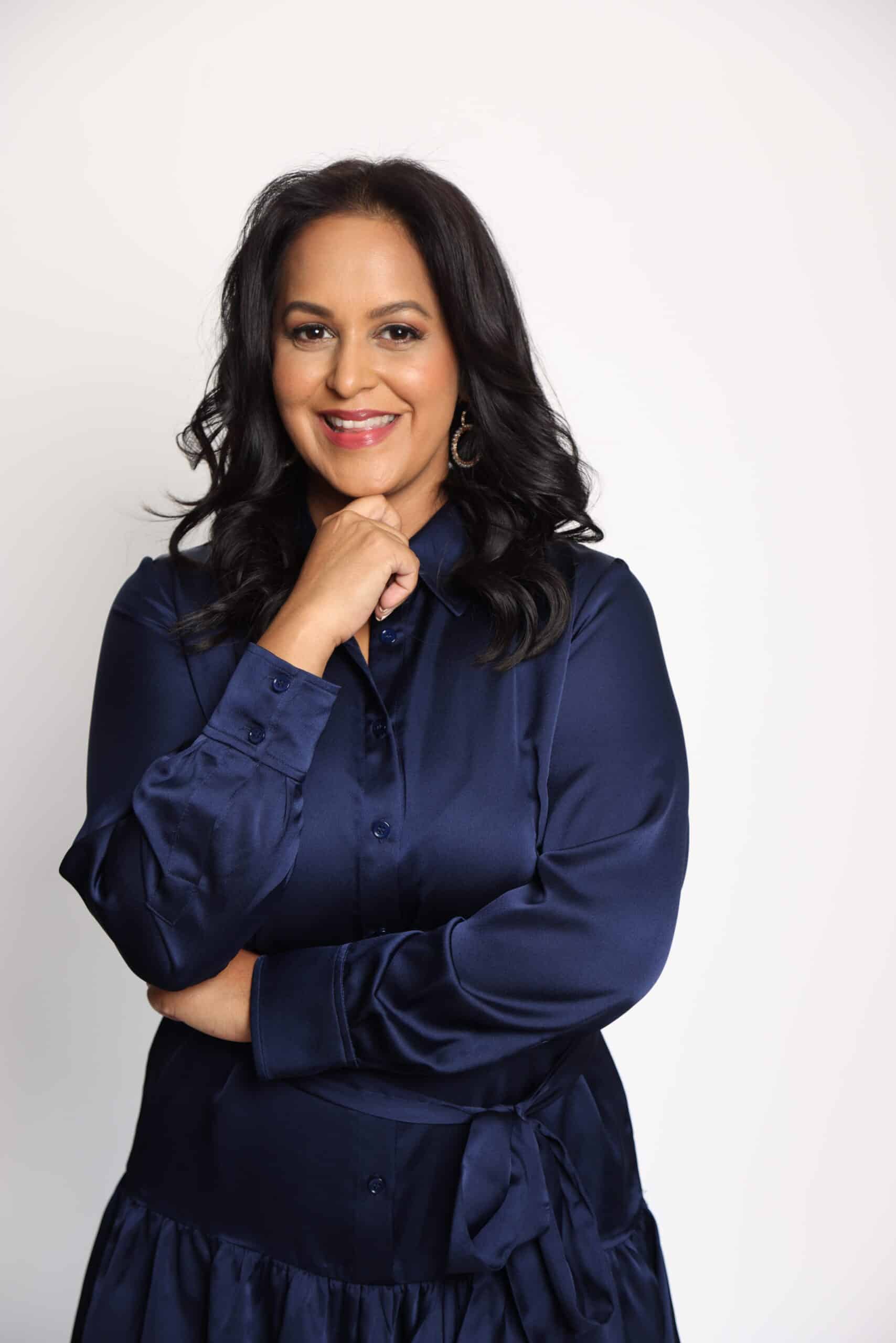 Smiling woman with long dark hair wearing a navy satin blouse, hand resting under her chin against a light background.